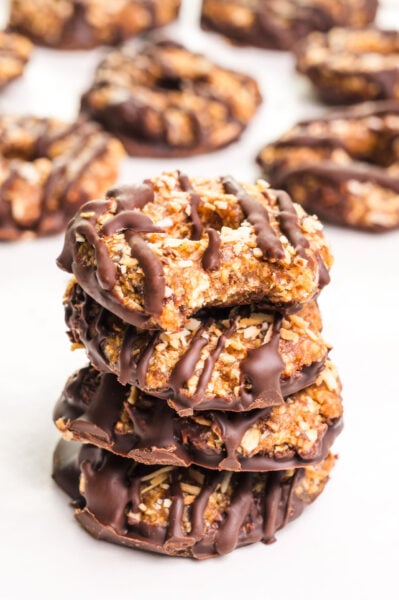 A stack of vegan Samoas cookies with a bite taken out of the top one sits in front of more cookies in the background.