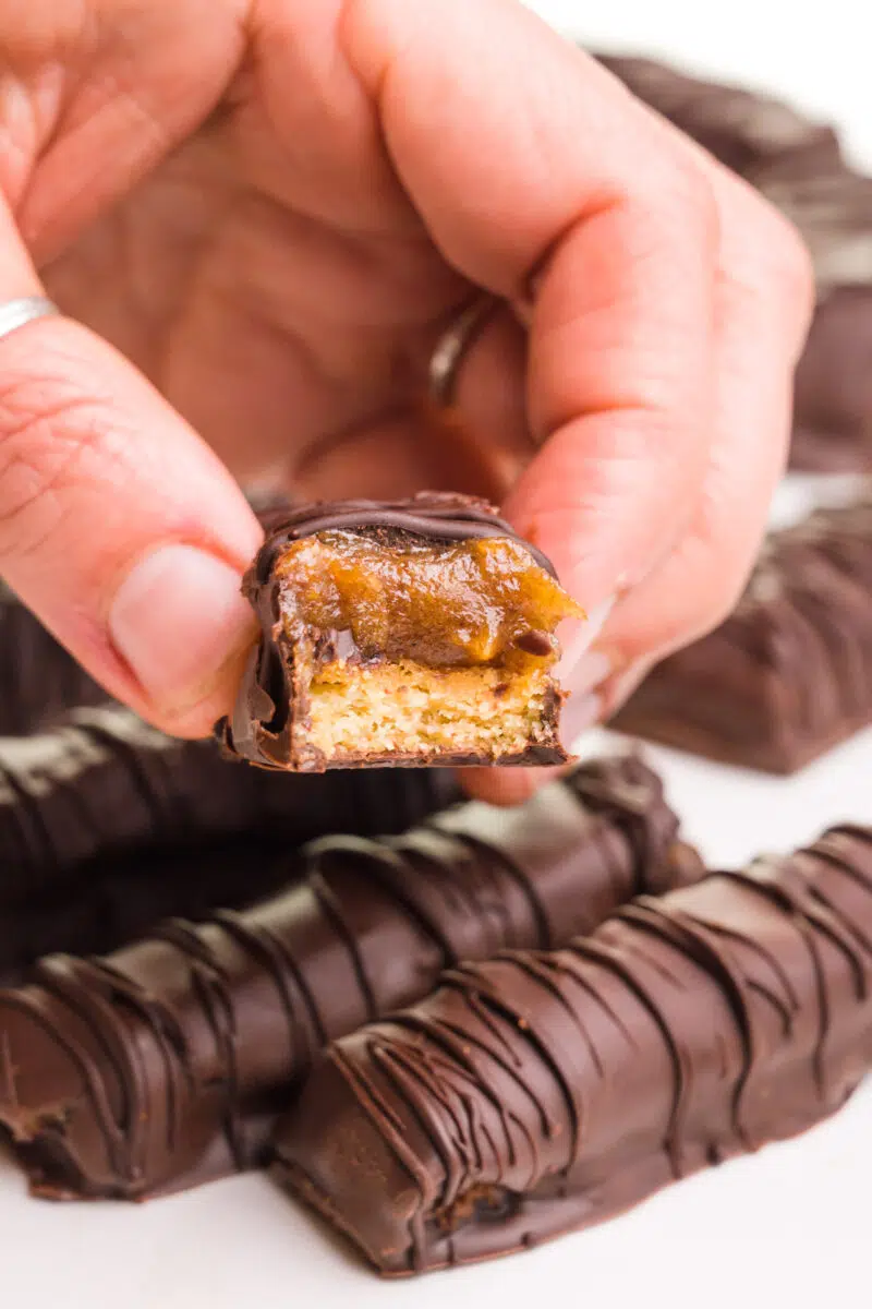 A hand holds a vegan Twix with a bite taken out. It hovers over more Twix bars.