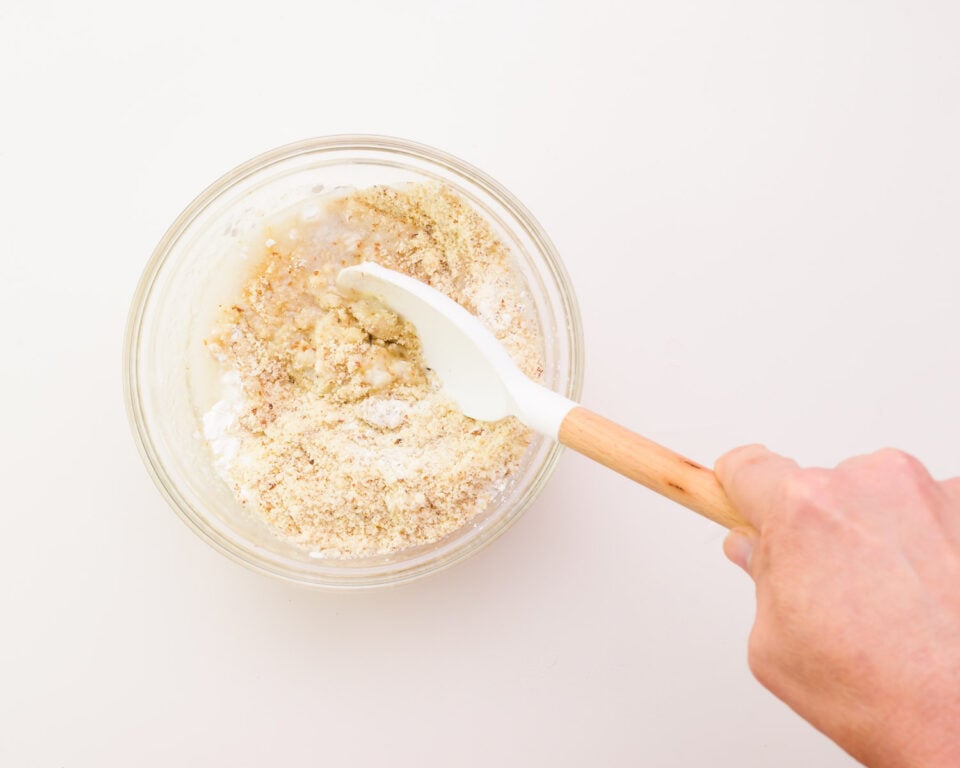 A hand holds a spatula, stirring flour ingredients in a bowl.
