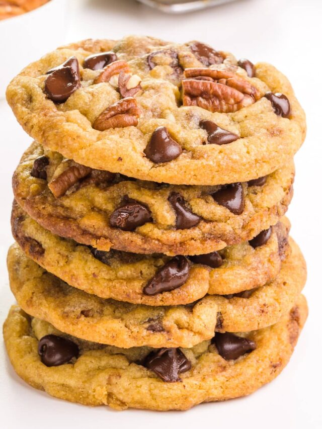 A stack of chocolate chip pecan cookies sits in front of a bowl of pecans and more cookies in the background.