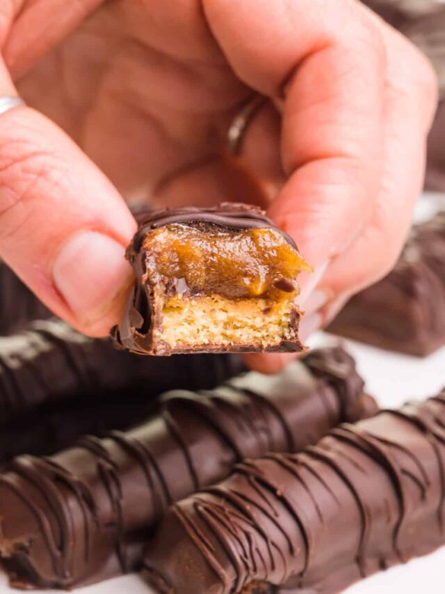 A hand holds a vegan Twix with a bite taken out. It hovers over more Twix bars.