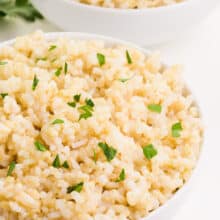 Two bowls of cooked brown rice sit next to sprigs of parsley.