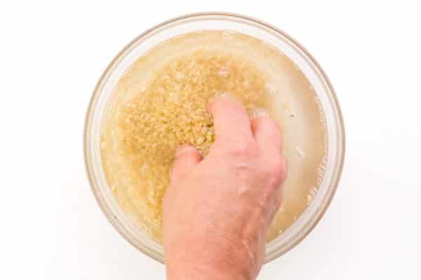 A hand reaches into a bowl with uncooked rice and water, stirring the rice.