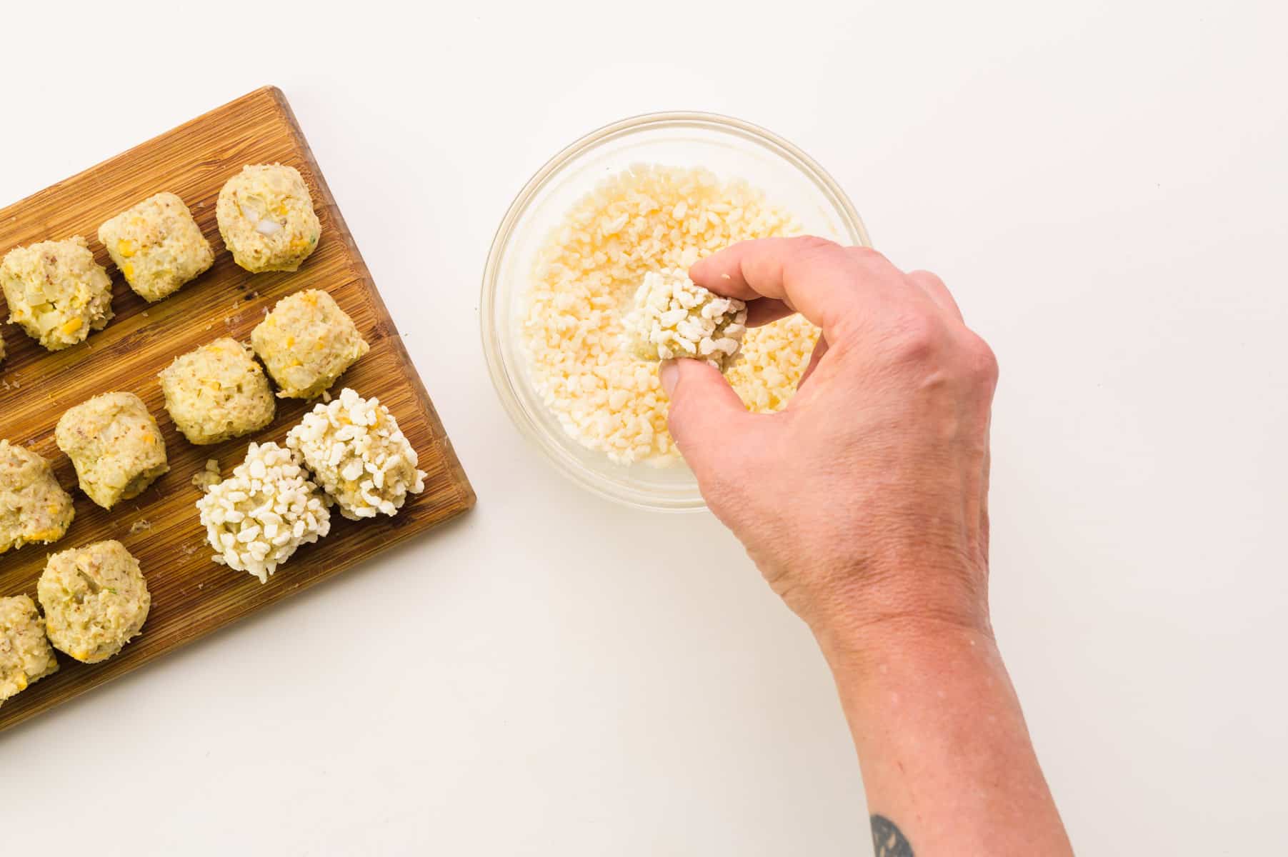 A hand rolls a cauliflower tot in bread crumbs. There is a cutting board with tots on the side.