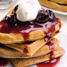 Waffles are smothered in cherry sauce and a dollop of whipped cream. There are more waffles and fresh cherries in the background.