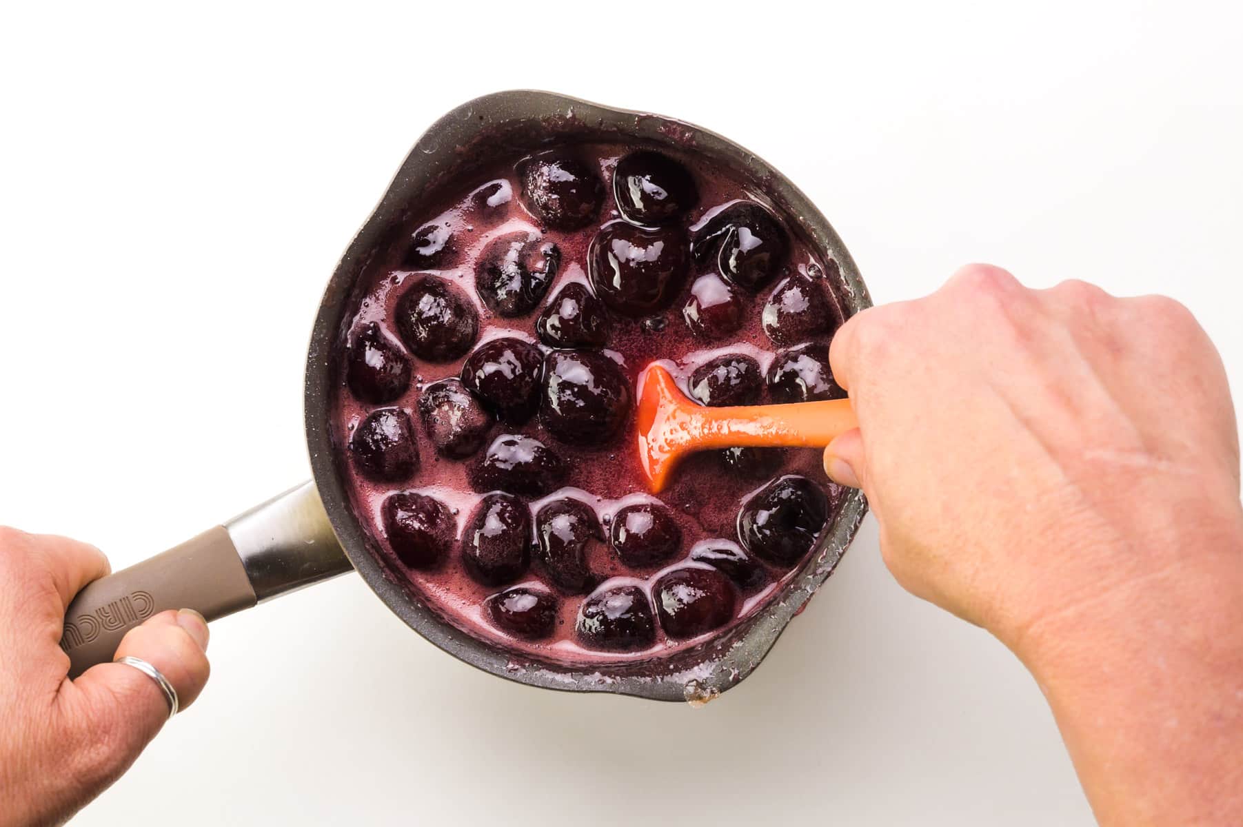 A hand holds a spatula, stirring cherry sauce in a saucepan.