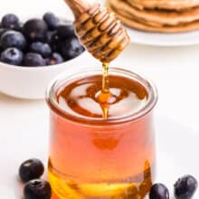A wooden honey dipper pours over a jar full of honey substitute.  In the background are fresh blueberries and pancakes.