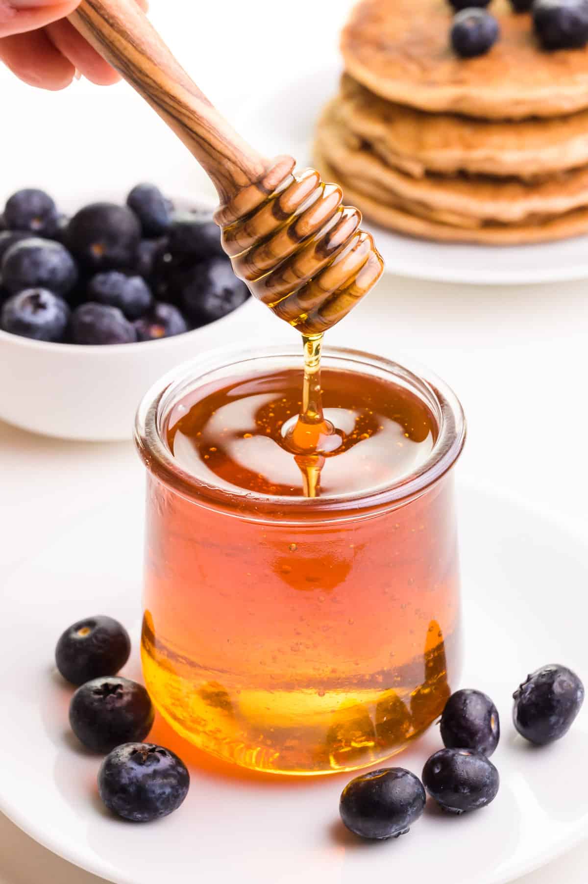 A wooden honey dipper drizzles over a jar full of honey substitute. There are fresh blueberries and pancakes in the background.
