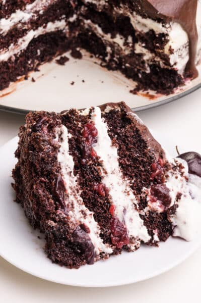 A slice of vegan black forest cake sits on a plate in front of the rest of the cake.