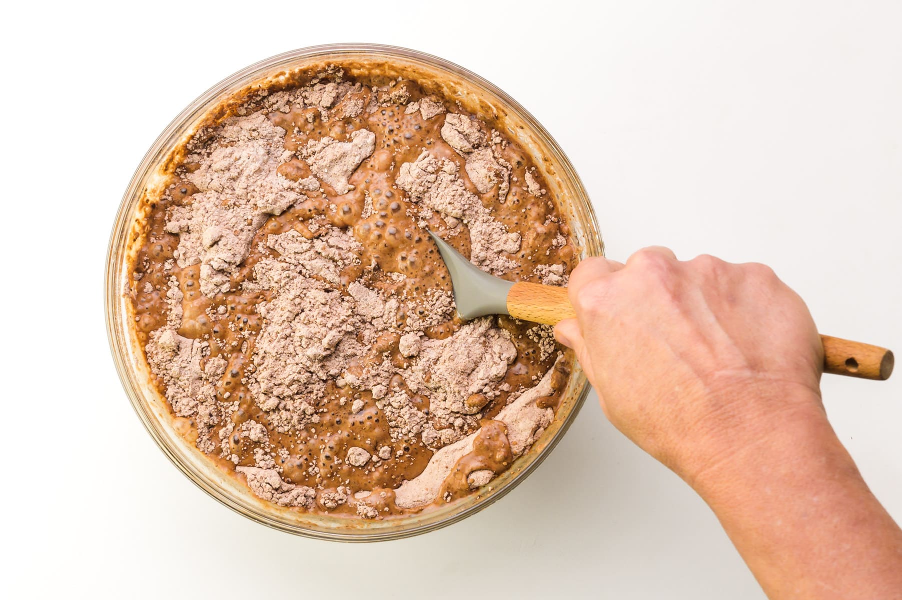 A hand holds a spatula, stirring cake batter in a bowl.
