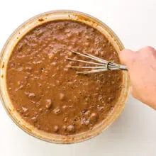 A hand holds a wire whisk, stirring chocolate cake batter in a bowl.
