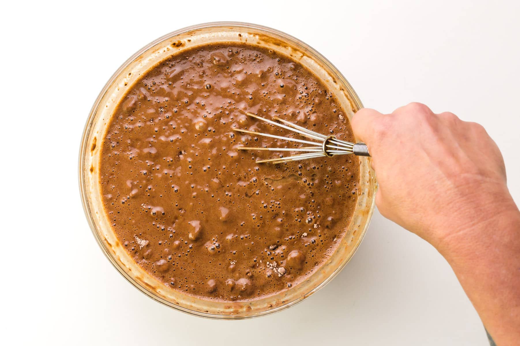A hand holds a wire whisk, stirring chocolate cake batter in a bowl.
