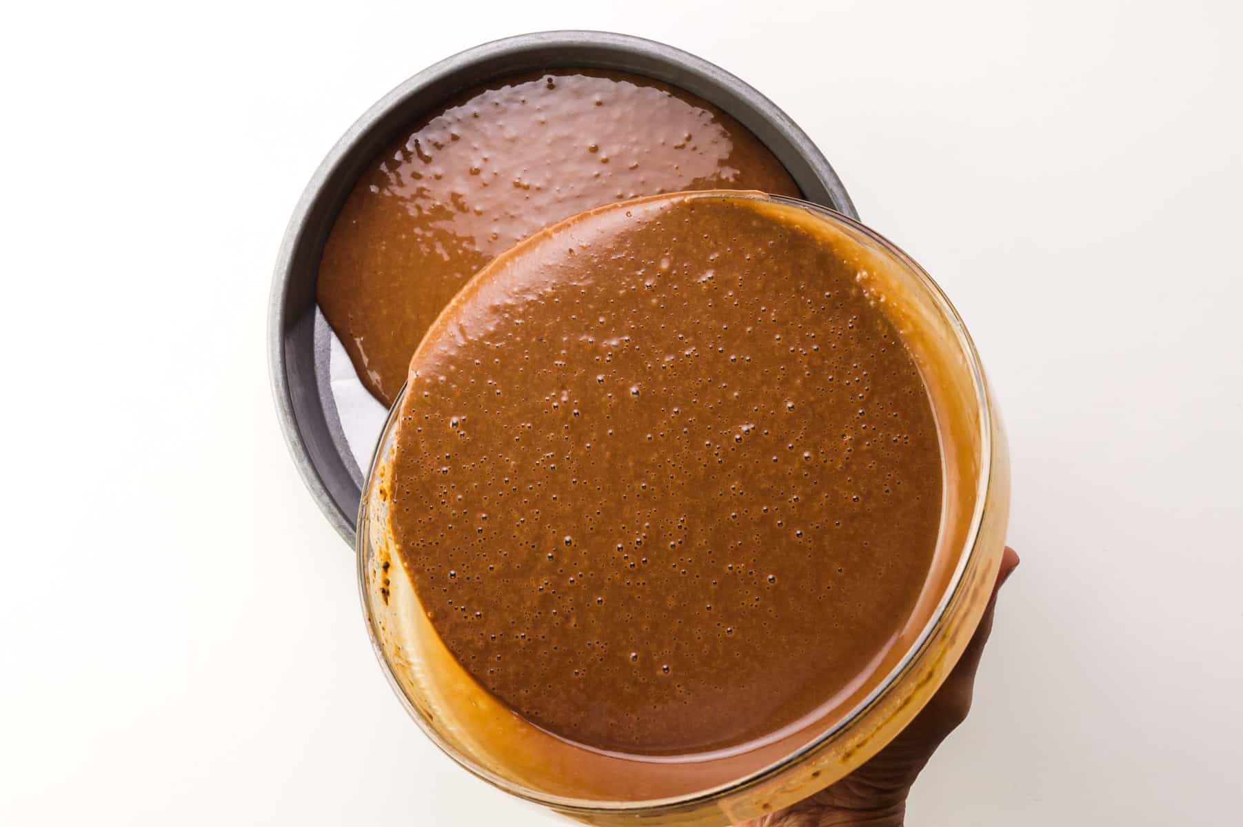 A hand holds a bowl of chocolate cake batter, pouring it into a prepared round cake pan.