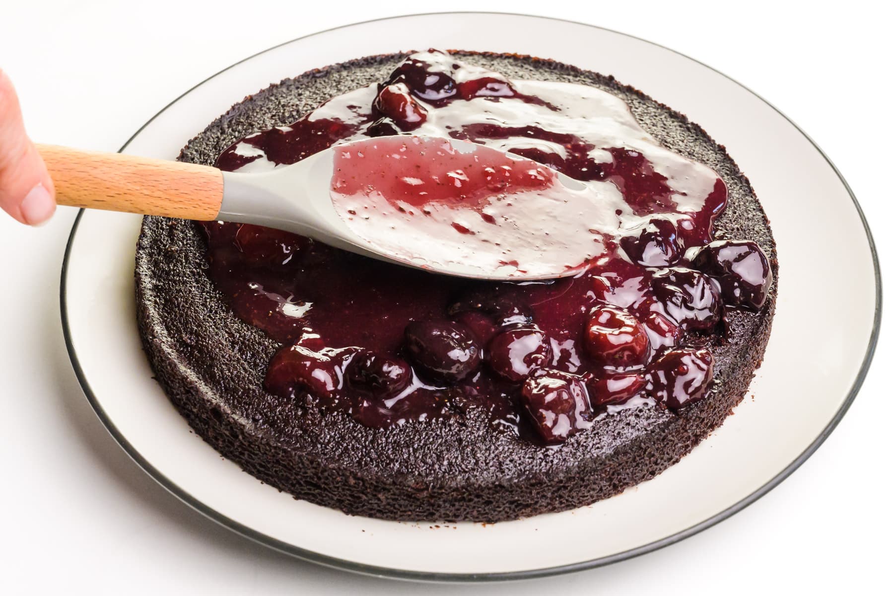 Cherry sauce is being spread with a spatula over a single chocolate cake layer.