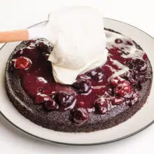 A spatula drops a dollop of whipped cream over cherry sauce on a chocolate cake.