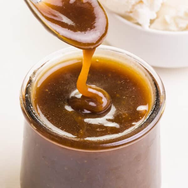 A spoon drizzles dairy-free butterscotch sauce over a jar with more sauce in it.
