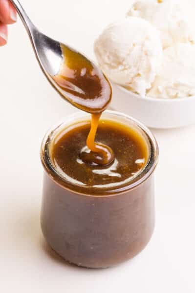 A hand holds a spoon with vegan butterscotch sauce drizzling over a glass jar full of the sauce. There is a bowl of ice cream in the background.