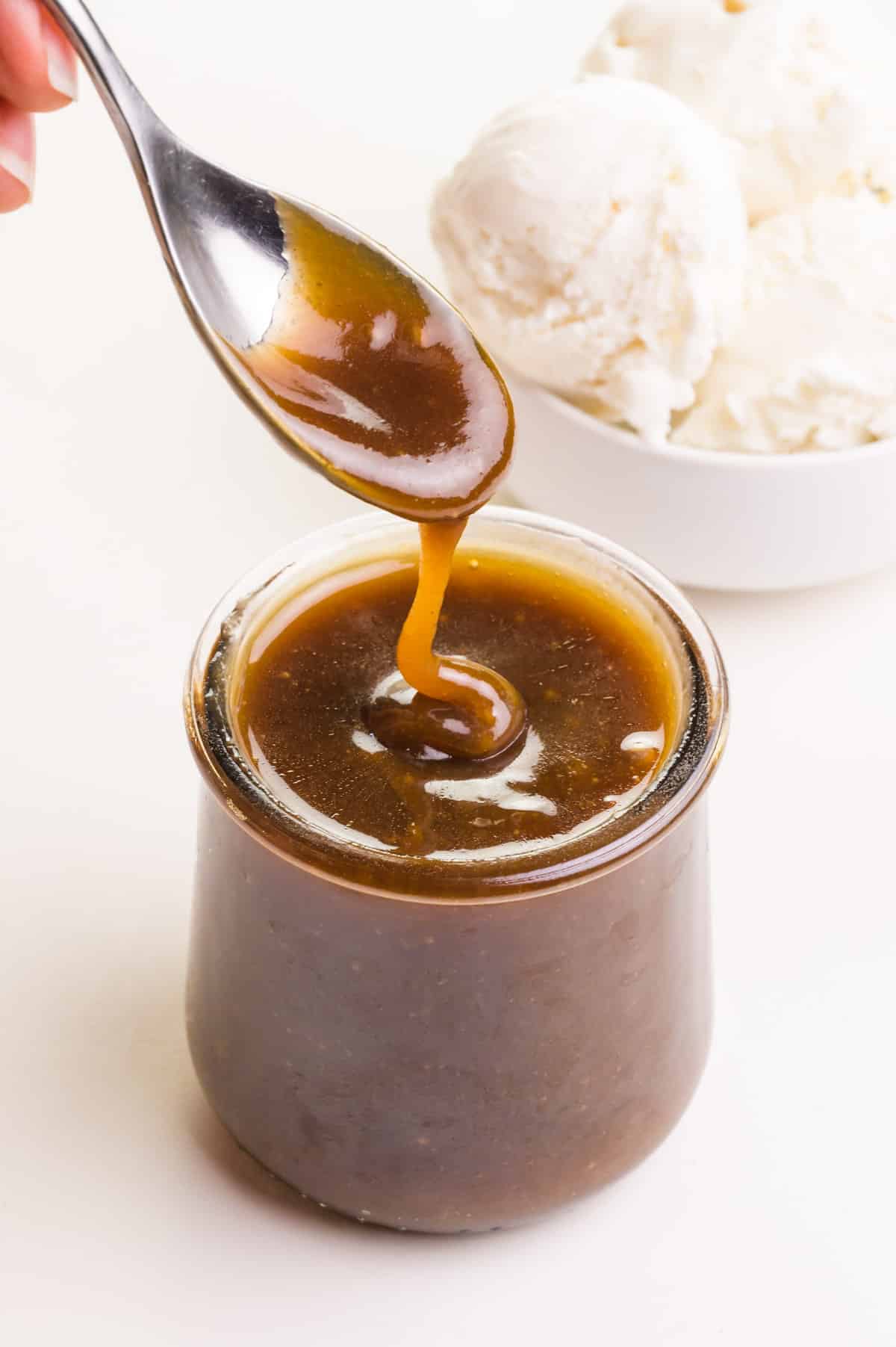 A hand holding a spoon with vegan butterscotch sauce on top of a glass jar full of sauce.  There is a bowl of ice cream in the background.