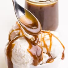 A spoon drizzles vegan butterscotch sauce over ice cream. There is a jar with more sauce in the background.