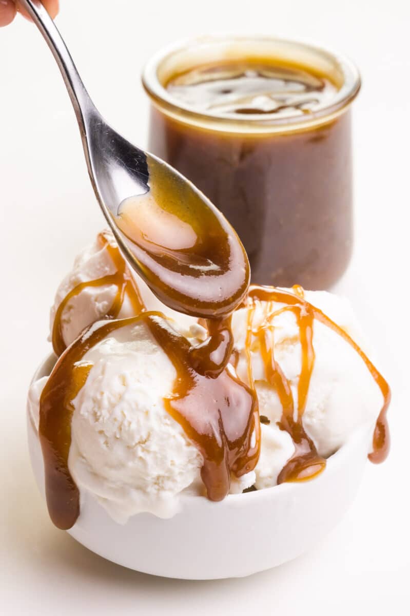 A spoon drizzles vegan butterscotch sauce over ice cream. There is a jar with more sauce in the background.