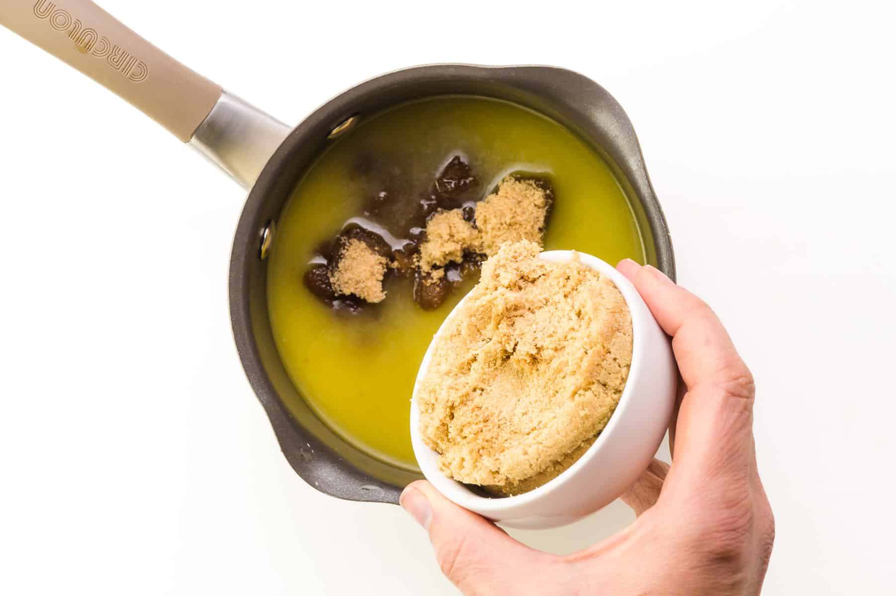 A hand holds a bowl of brown sugar, pouring it into a saucepan with melted butter.