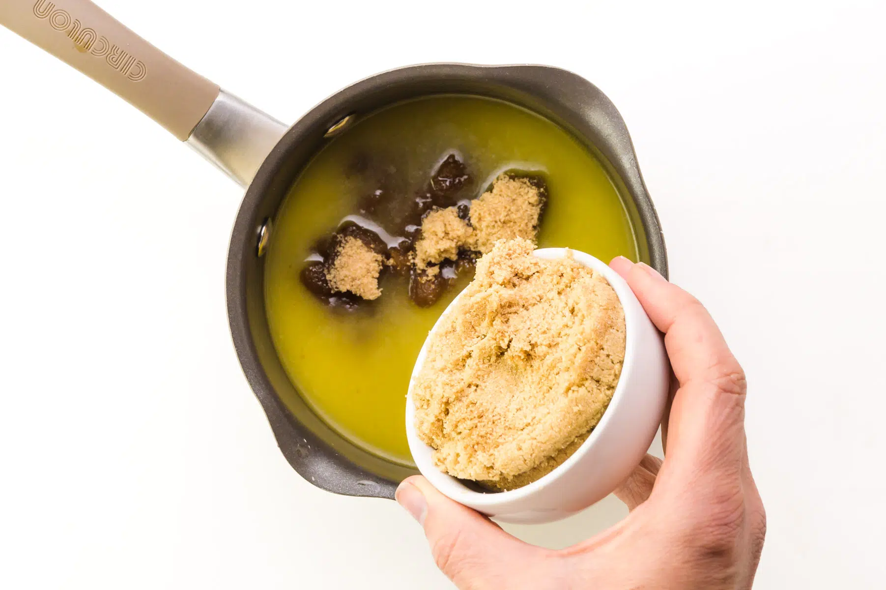 A hand holds a bowl of brown sugar, pouring it into a saucepan with melted butter.