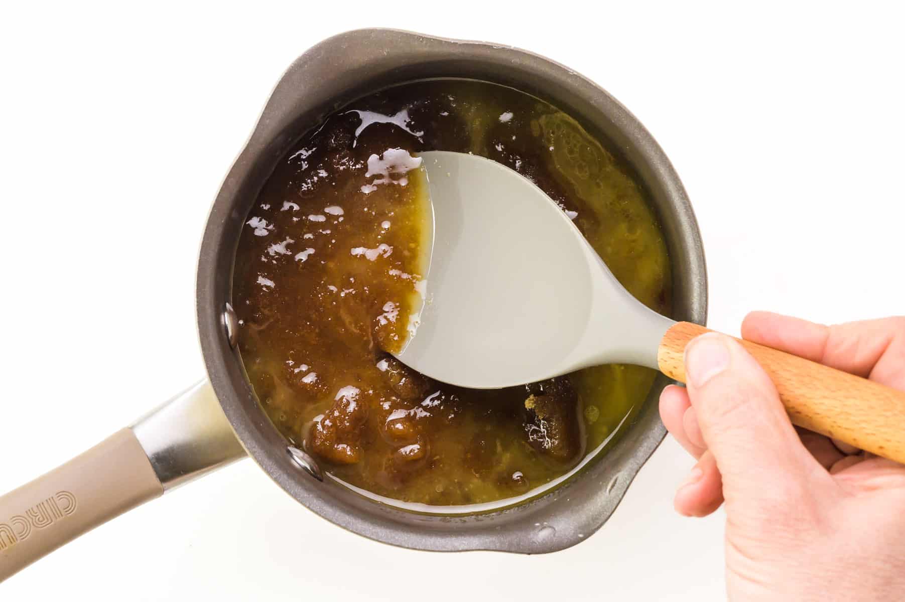 A hand holds a spatula, stirring brown sugar syrup in a saucepan.