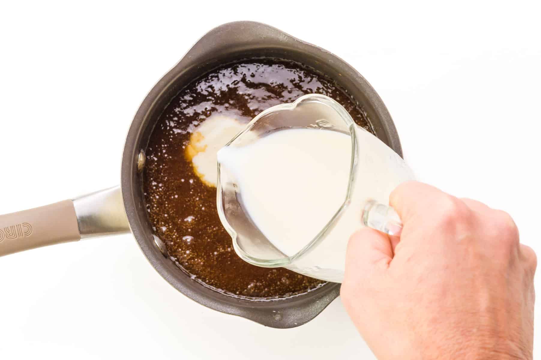 A hand holds a small pitcher of plant-based milk, pouring it into a saucepan full of brown sugar syrup.