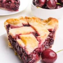 A slice of vegan cherry pie sits on a plate with a fresh cherry next to it. There is a bowl of cherries and another slice of pie in the background.
