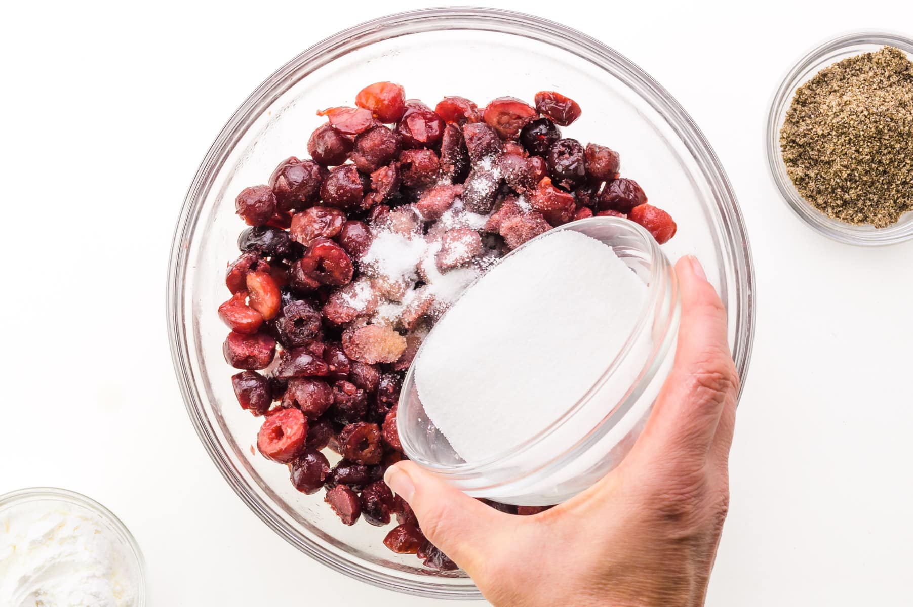 A hand holds a bowl of sugar, pouring it into a bowl of cherries. There is a bowl of ground flax seed nearby.