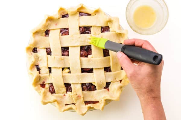 A hand holds a pastry brush, brushing vegan egg wash over a lattice pie. There is a bowl with yellow liquid on the top right.