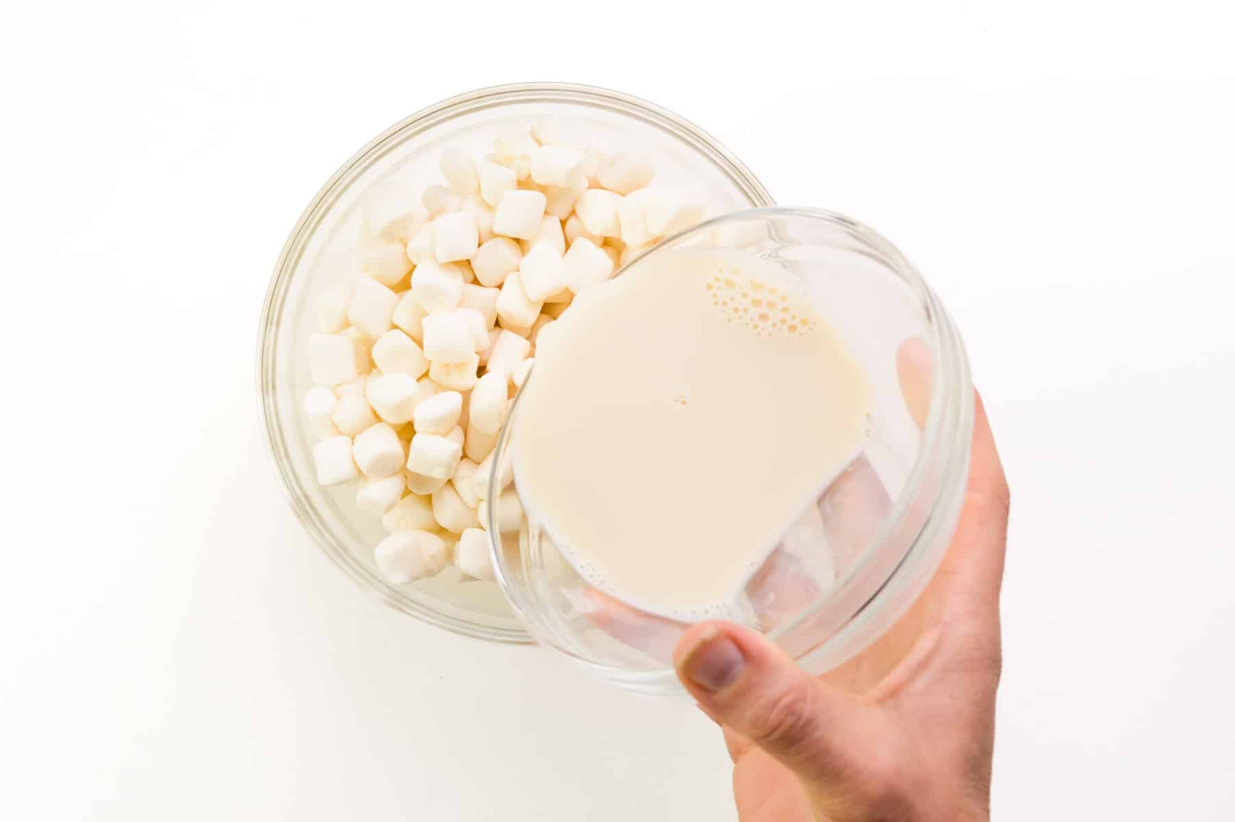 A hand holds a bowl of soy milk, pouring it into a bowl of marshmallows.