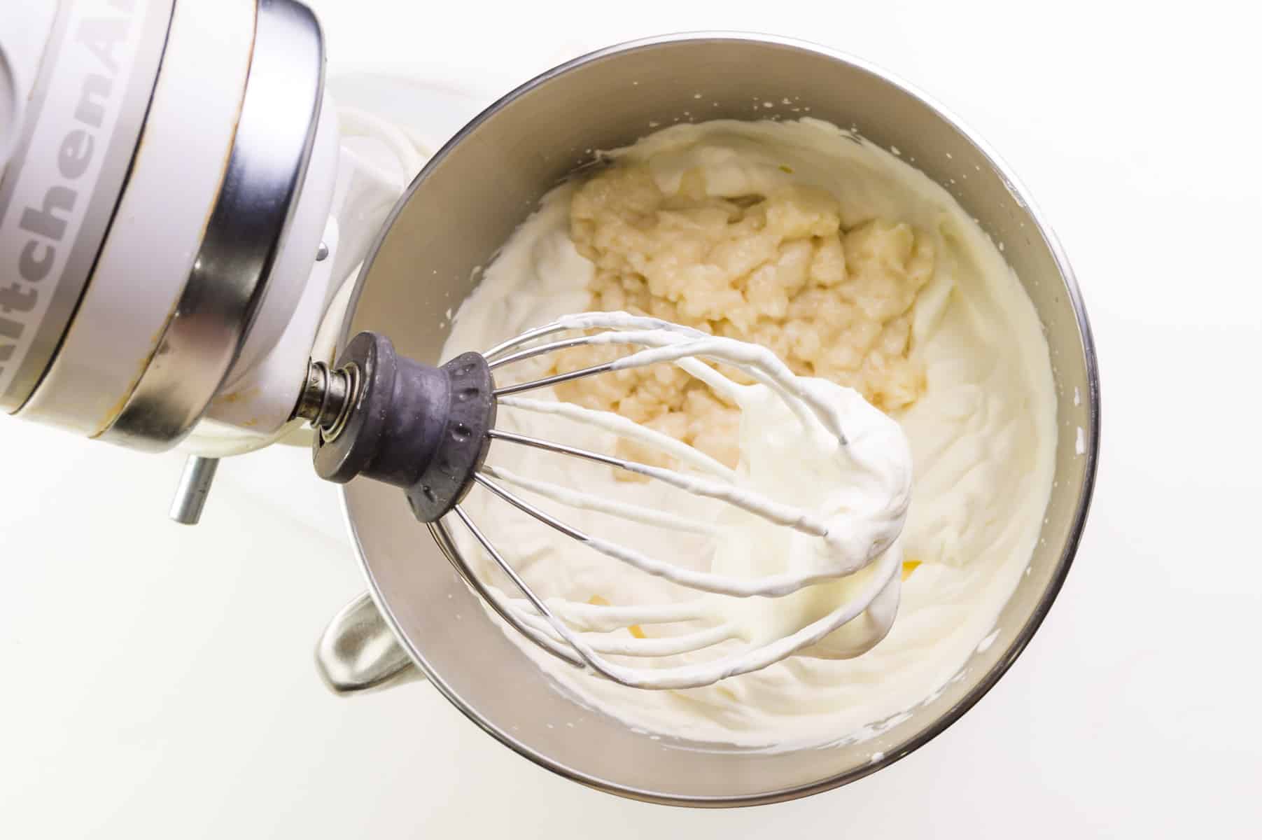 A marshmallow mixture is added to the whipped cream in the mixing bowl of a stand mixer.