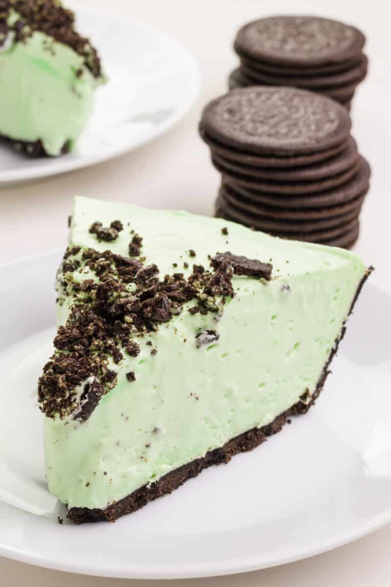 A slice of vegan grasshopper pie sits in front of a stack of sandwich cookies and another slice of pie.