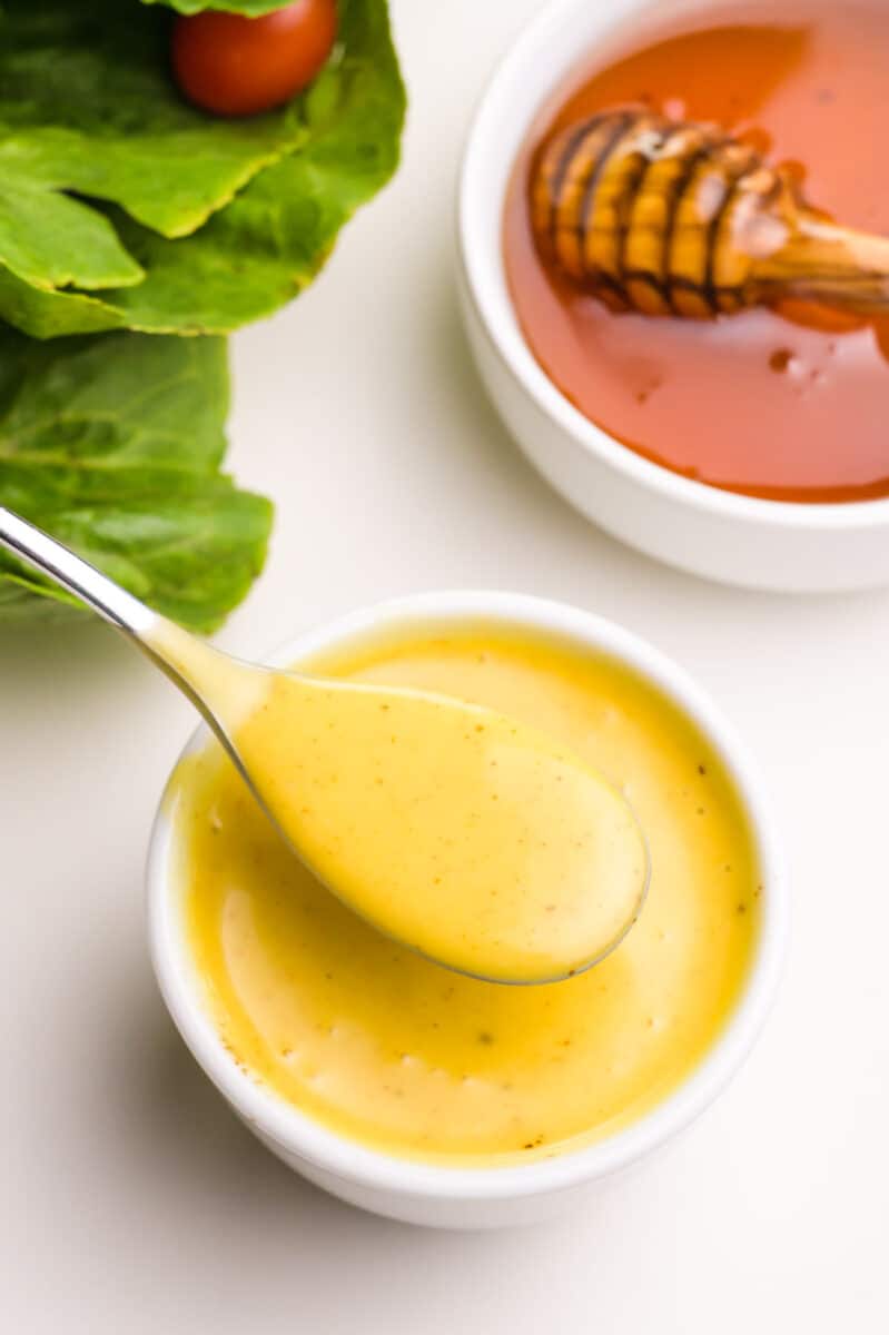 Looking down on a bowl of dairy-free honey mustard with a spoon in it. There are greens in the background and a bowl of honey with a honey stick in it.