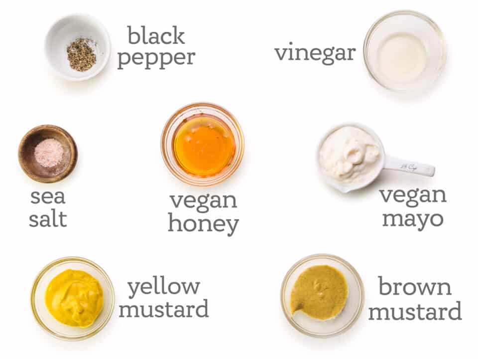Ingredients are laid out on a white table. The labels next to them read, vinegar, vegan mayo, brown mustard, yellow mustard, vegan honey, sea salt, and black pepper.