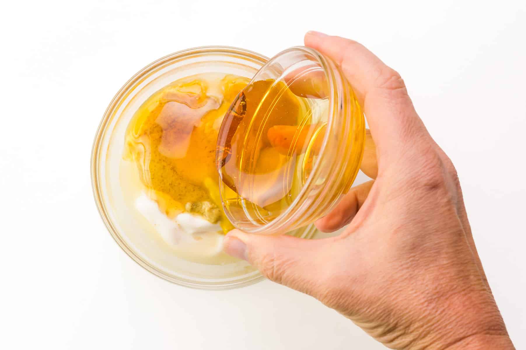A hand holds a bowl of honey, pouring it into a bowl with mayo and mustard.