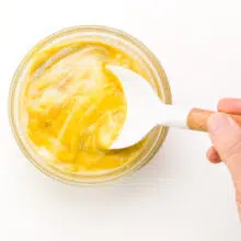 A hand holds a spatula stirring a small bowl of mustard and honey.