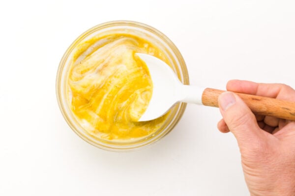 A hand holds a spatula stirring a small bowl of mustard and honey.