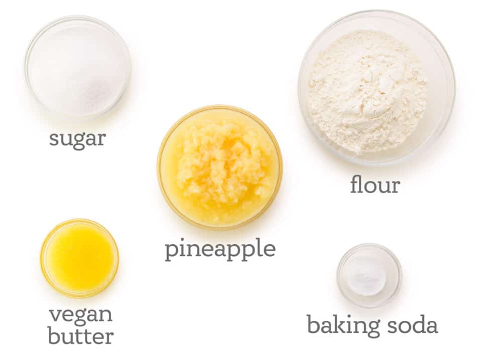 Ingredients are laid out on a white counter. The labels next to them read, flour, baking soda, pineapple, vegan butter, and sugar.
