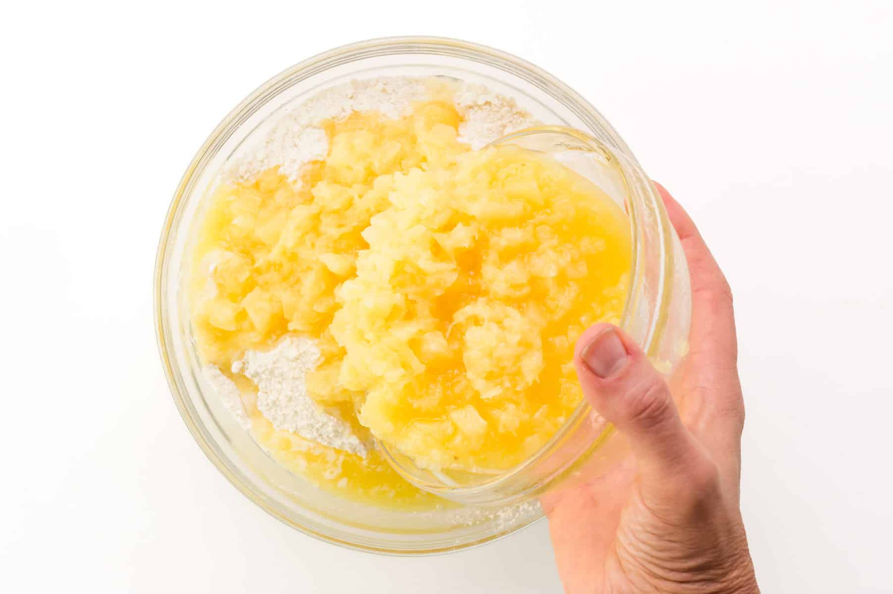 A hand holds a bowl with crushed pineapple, pouring into a bowl with flour.