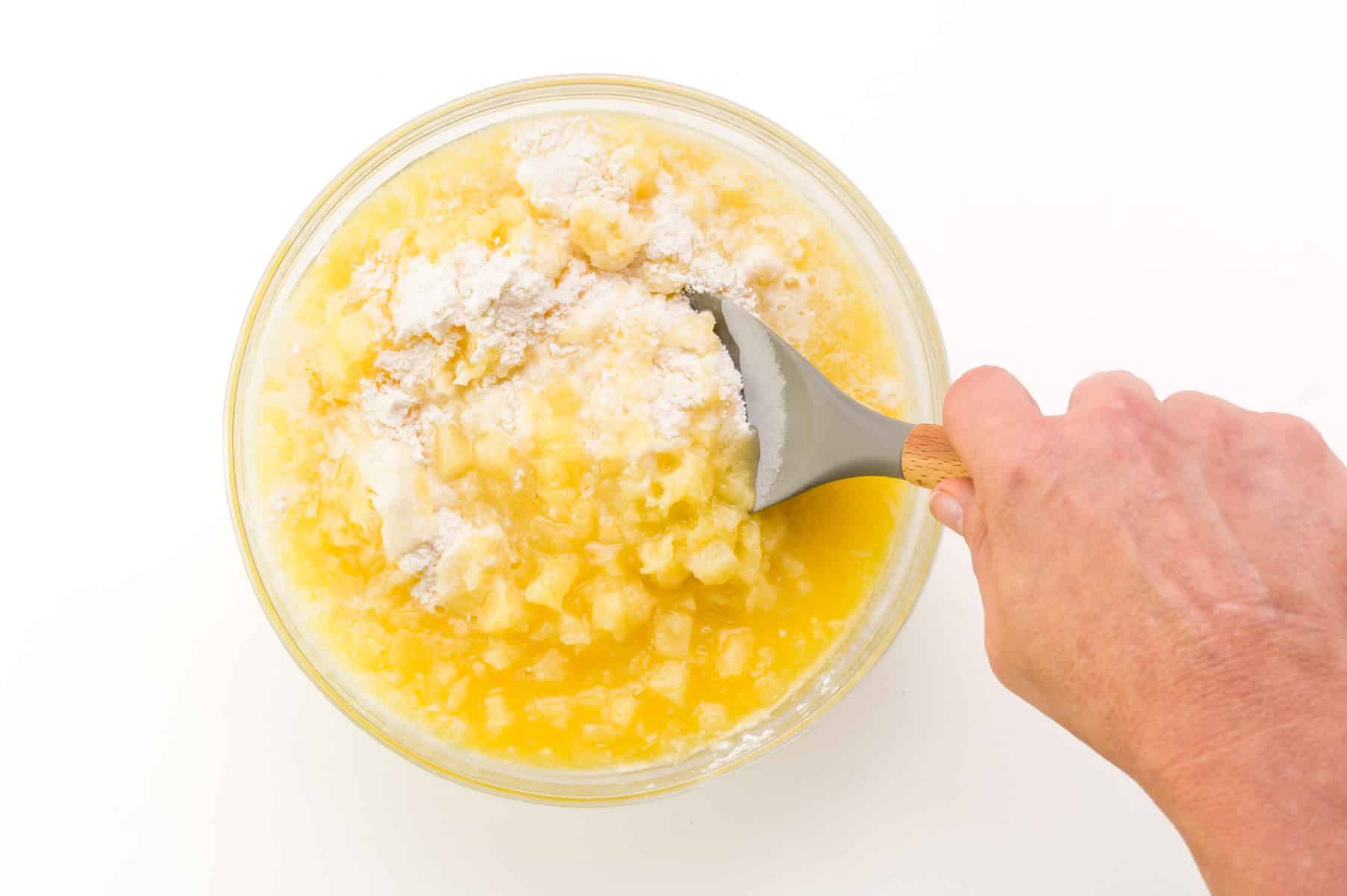 A hand holds a spatula, using it to stir together cake batter with crushed pineapple.