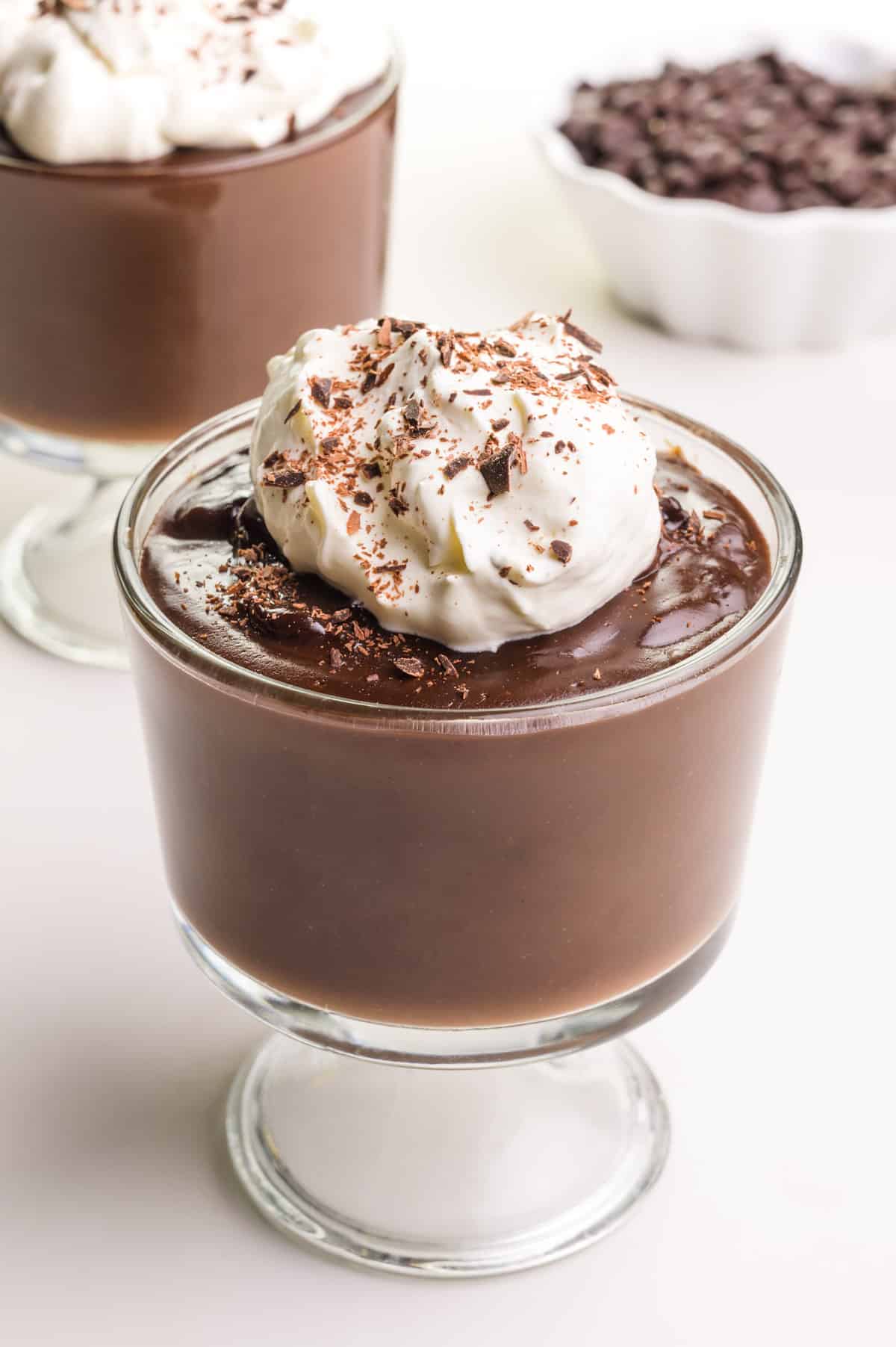A bowl of vegan chocolate pudding has whipped cream on top. It sits in front of another bowl of pudding and a bowl of chocolate chips.