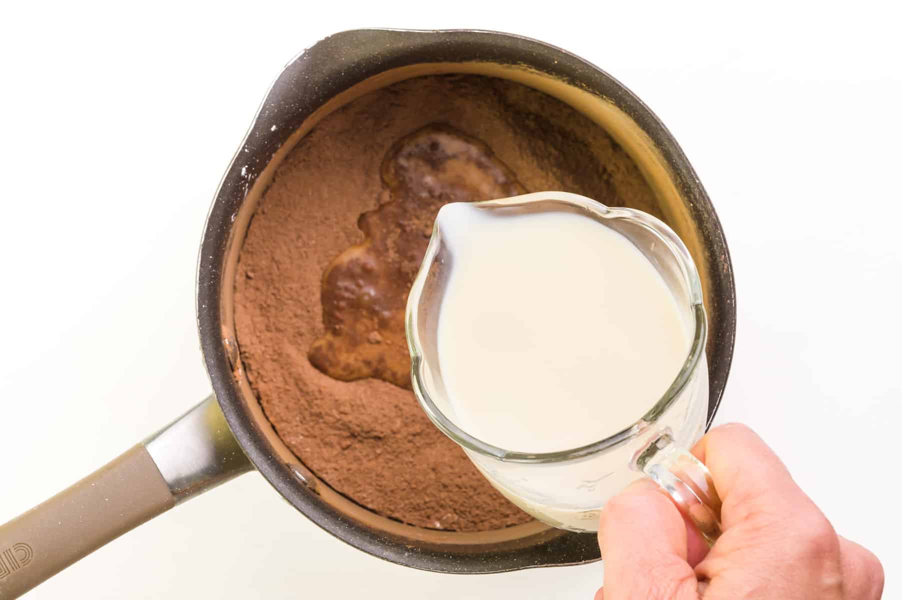 Milk is being poured into a saucepan with a cocoa powder mixture.