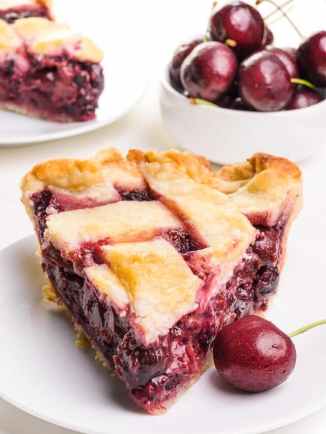 A slice of vegan cherry pie sits on a plate with a fresh cherry next to it. There is a bowl of cherries and another slice of pie in the background.
