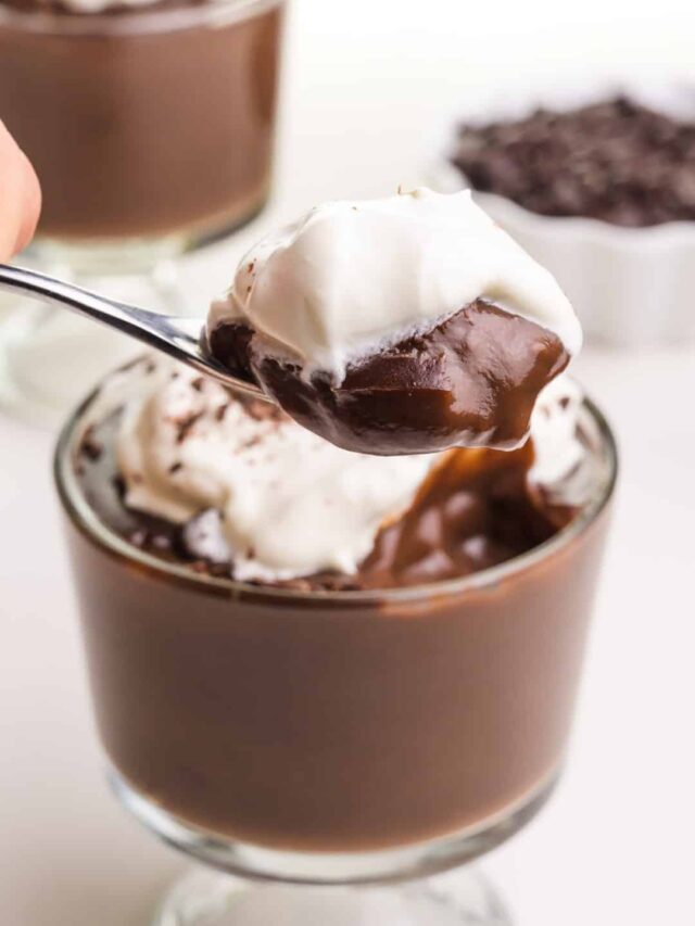 A hand holds a spoonful of vegan chocolate pudding topped with whipped cream. It hovers over a serving dish full of pudding.