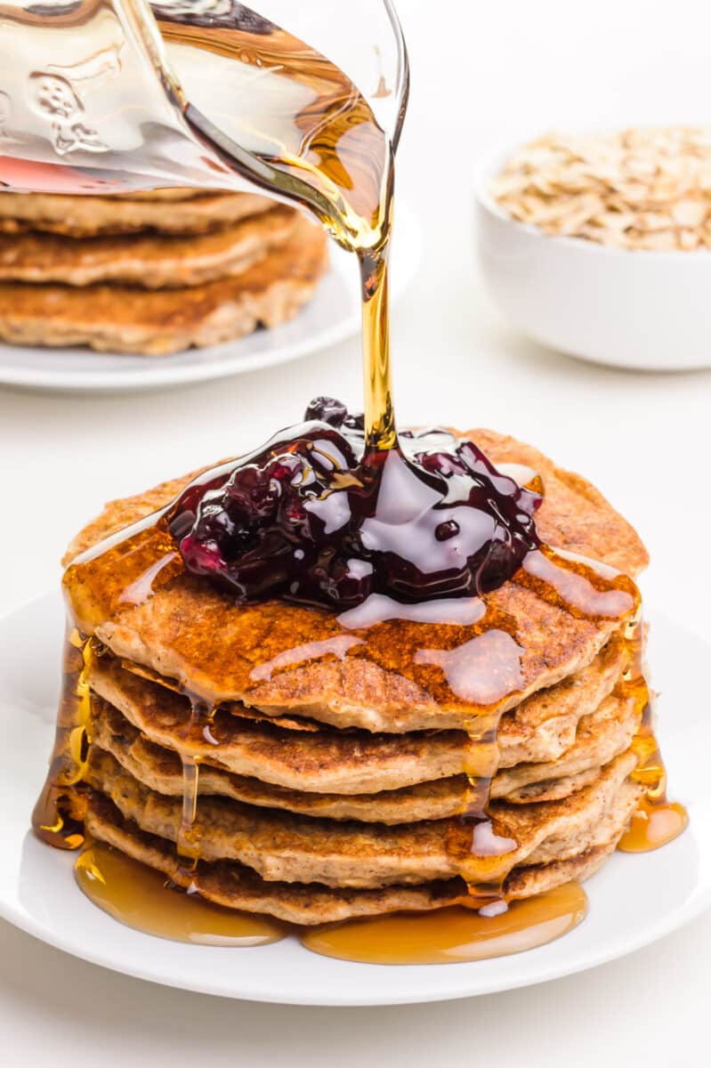 Vegan oatmeal pancakes are on a plate with blueberry sauce on top. Maple syrup is being poured on top. There is another plate of pancakes and a bowl of oats in the background.