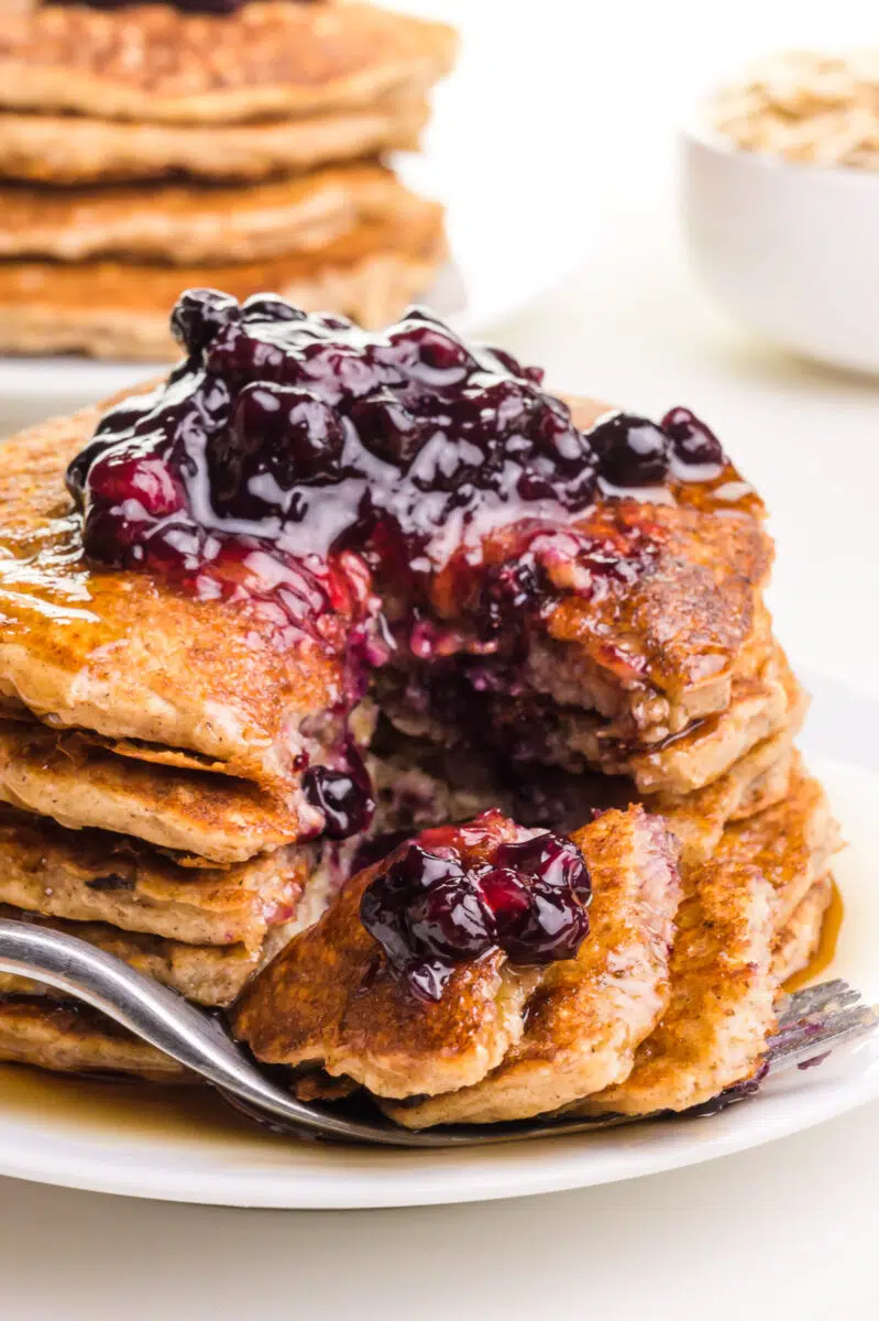 A fork full of cut pancake sits in front of a stack of pancakes with blueberries on top. There are pancakes in the background.