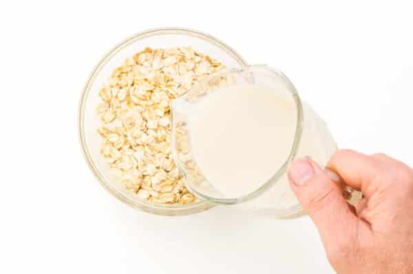 A hand holds a pitcher of milk, pouring it into a bowl of oats.