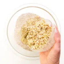 A hand holds a bowl of moistened oats, pouring them into a bowl with flour.